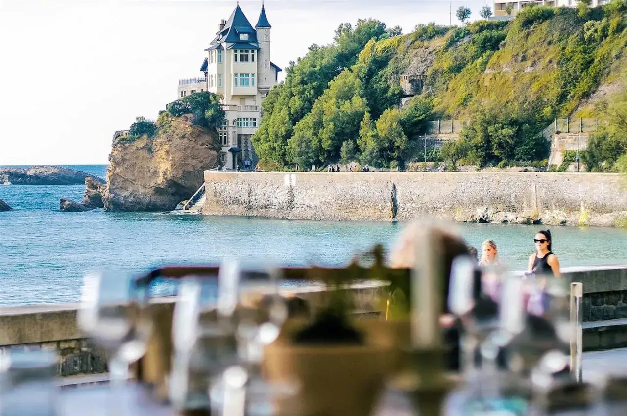Vue sur un château sur une falaise à Biarritz, avec la mer bleue et des verres en premier plan. Idéal pour savoir où manger à Biarritz.