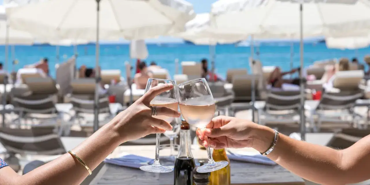 Deux mains portent des verres de rosé pour un toast, avec la mer bleue et des parasols en arrière-plan. Ambiance estivale dans un bar de plage à Cannes.
