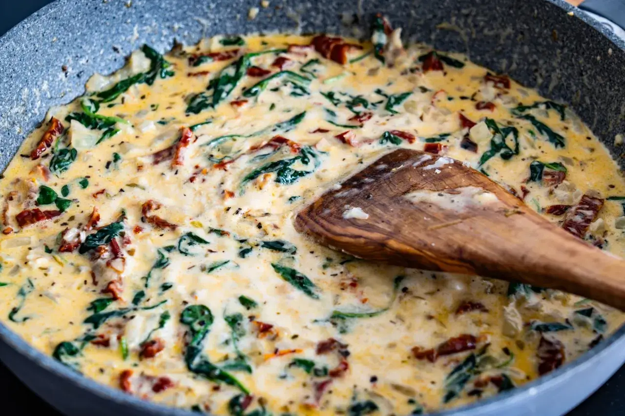 Une poêle de pâtes à la toscane crémeuses avec des épinards et des tomates séchées, mélangées avec une cuillère en bois.