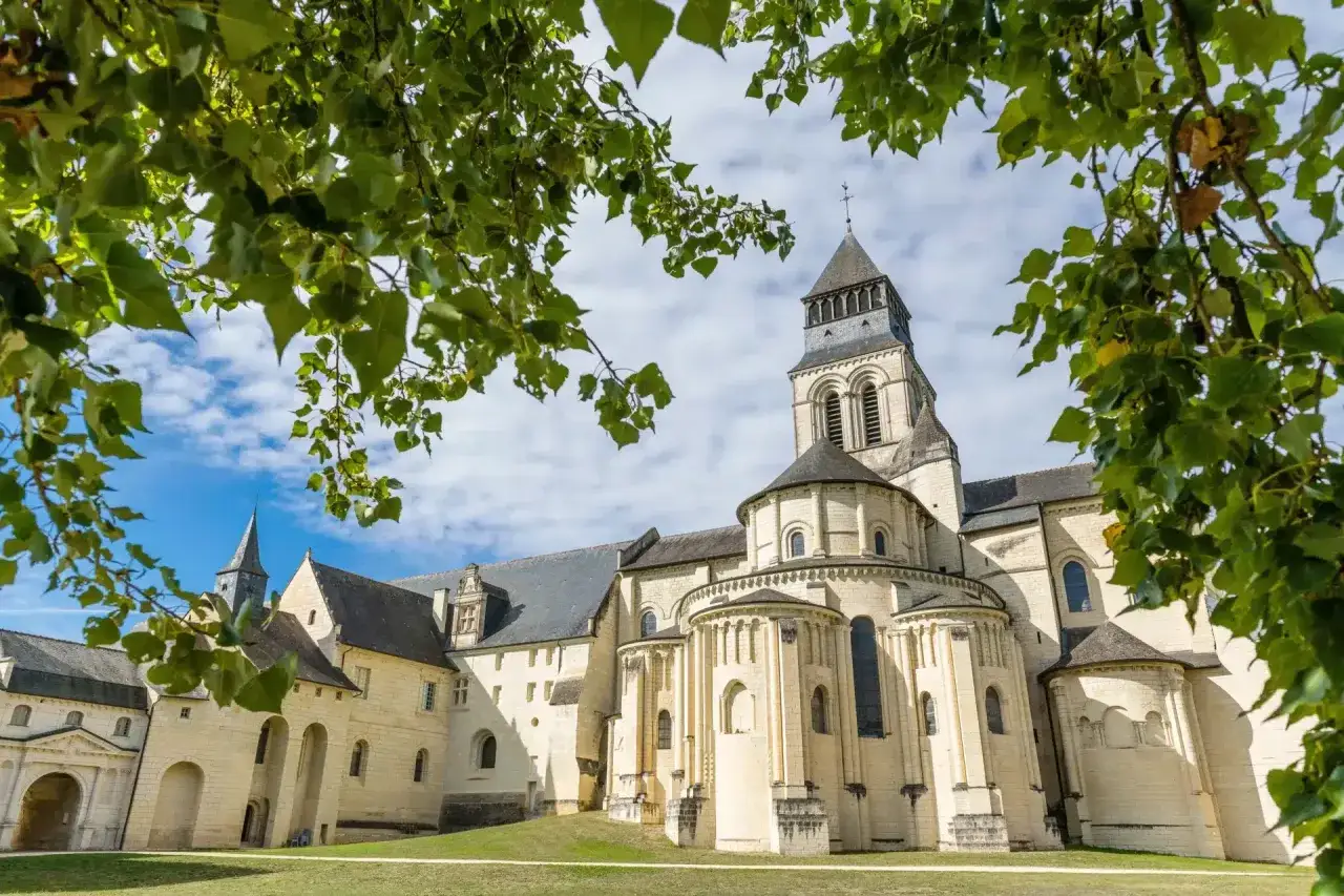 Le majestueux château de Fontevraud, avec sa tour imposante et ses absides romanes, se dresse sous un ciel bleu parsemé de nuages, encadré par le feuillage verdoyant des arbres.