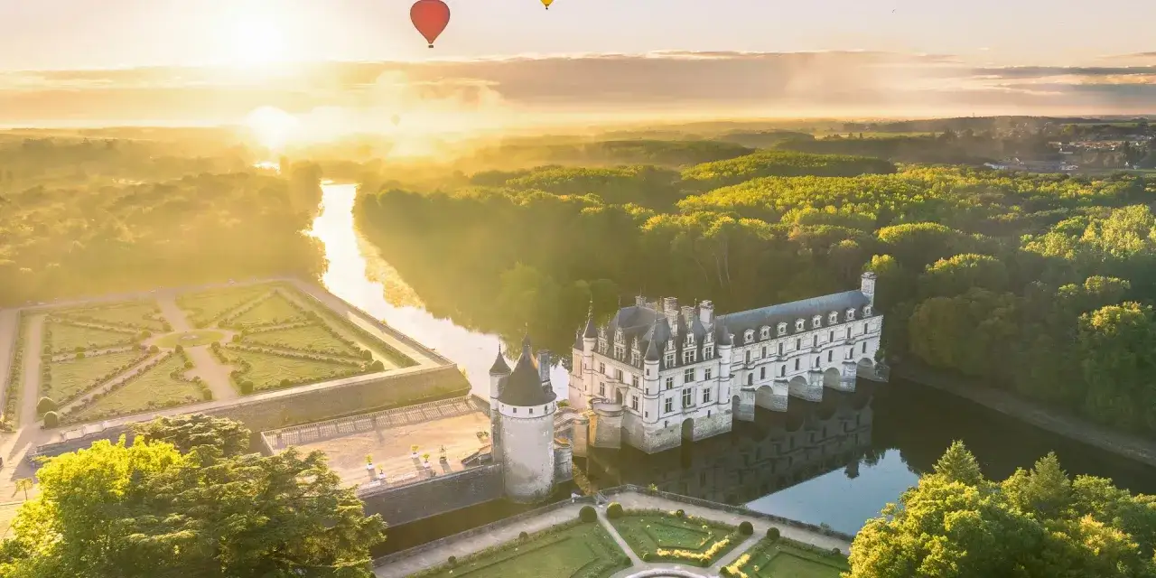 Lever du soleil sur les tours du château de Chenonceau, avec des montgolfières flottant au-dessus des jardins et de la rivière.