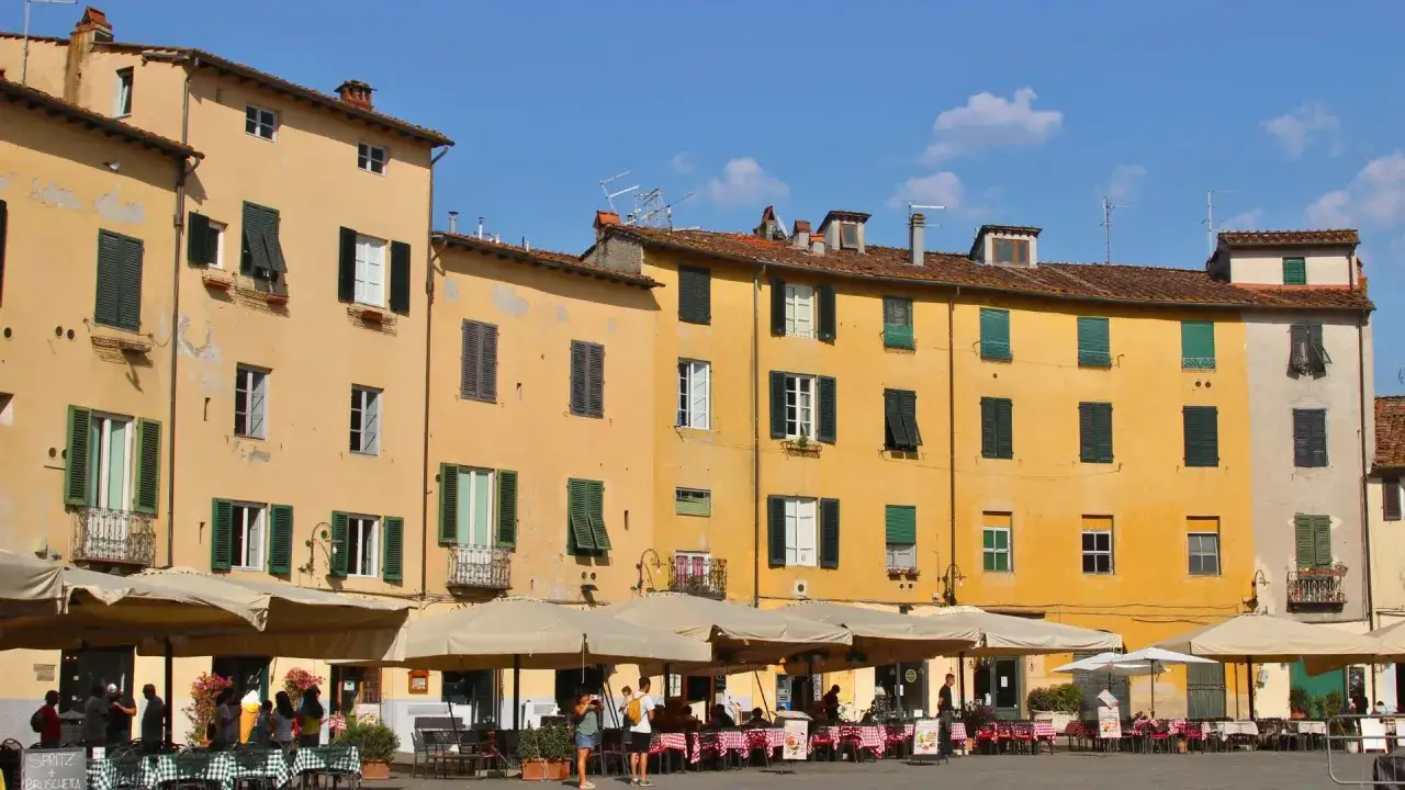 Terrasses animées sous le soleil de Lucca, Italie. Bâtiments ocre aux volets verts, tables dressées pour un repas en plein air.