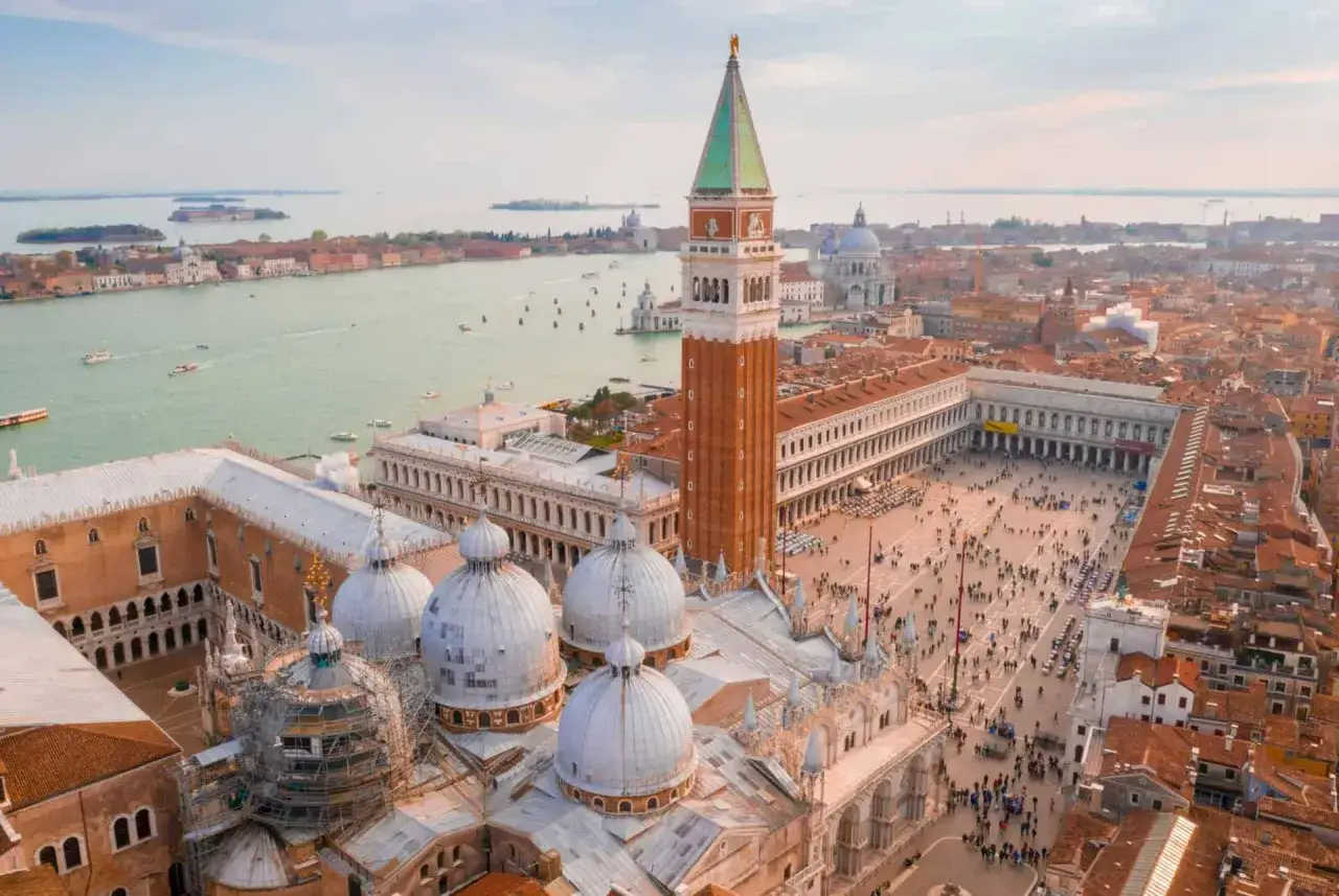 Vue aérienne de la Piazza San Marco à Venise, avec la basilique, le campanile et le canal. Une foule anime la place.