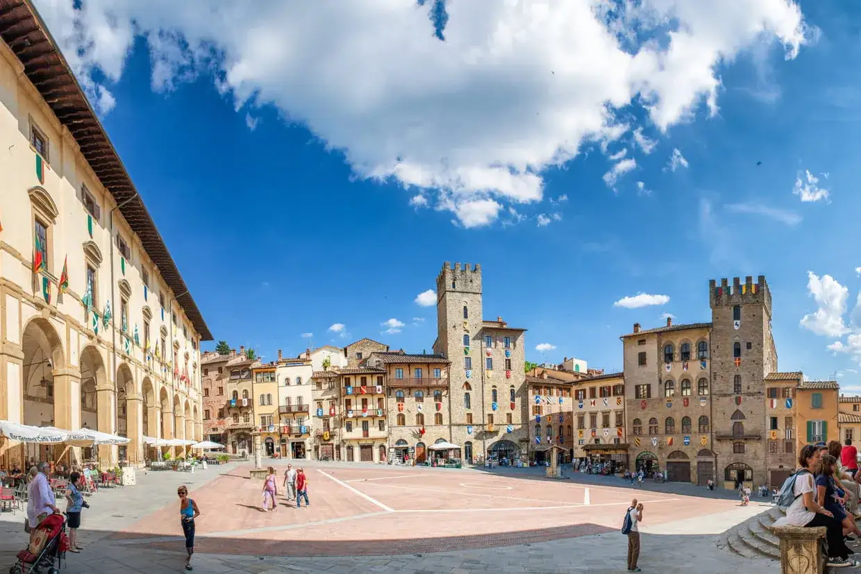 Arezzo, la vie est belle : une place anim&eacute;e avec des b&acirc;timents historiques, des tours m&eacute;di&eacute;vales et des drapeaux color&eacute;s sous un ciel bleu &eacute;clatant.