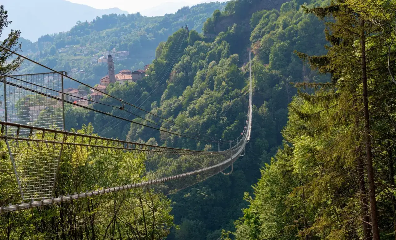Pont suspendu traversant une vallée verdoyante près de San Marcello Pistoiese, avec un village toscan en arrière-plan.