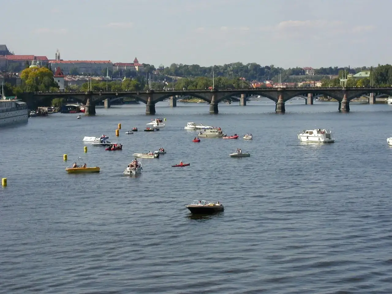Bateaux et canoës naviguent sur la Vltava, sous un pont, avec la ville de Prague en arrière-plan. Une scène de