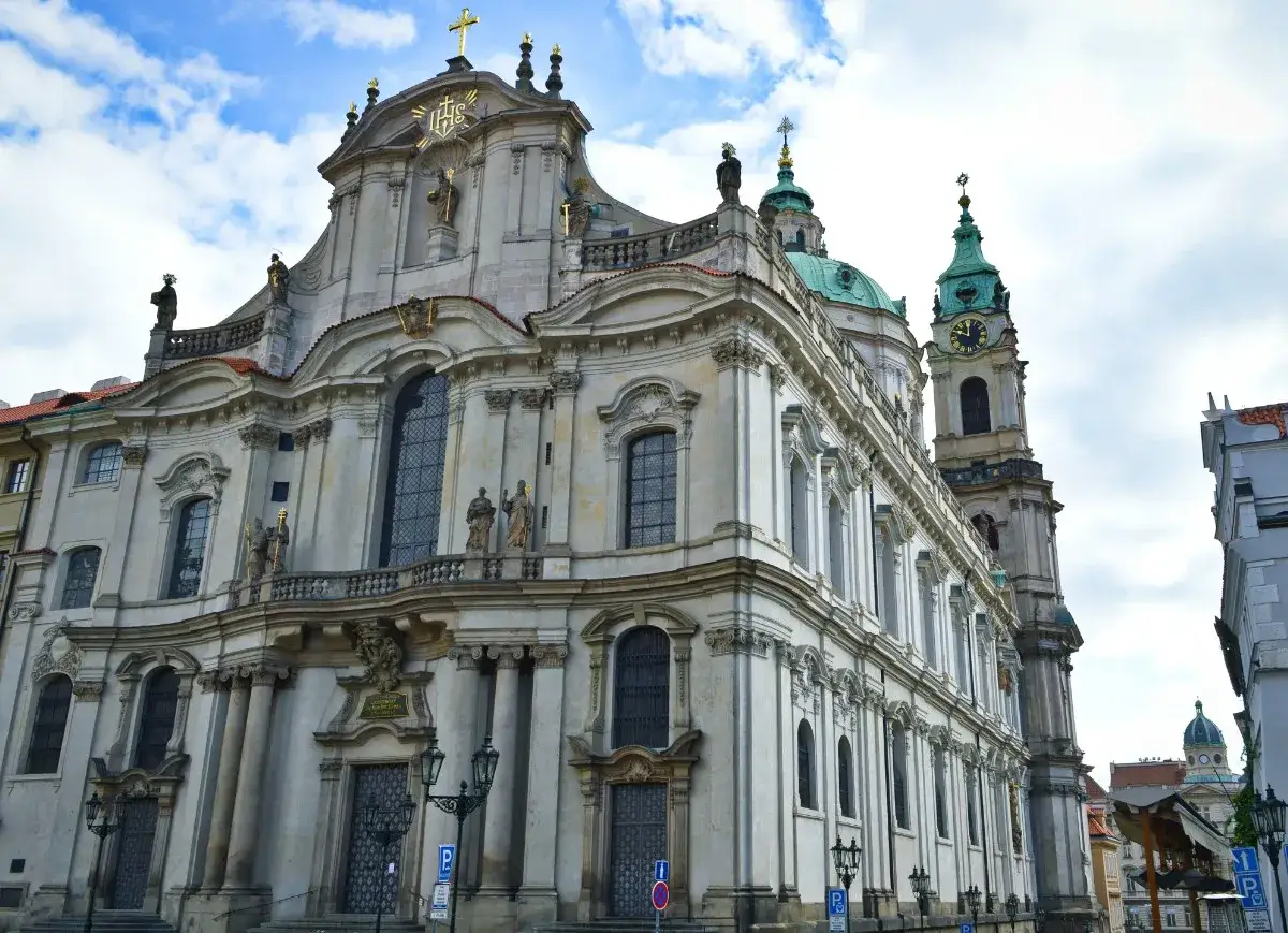 Magnifique façade de l'église Saint-Nicolas de Prague, avec sa tour à horloge et ses dômes verts sous un ciel nuageux.