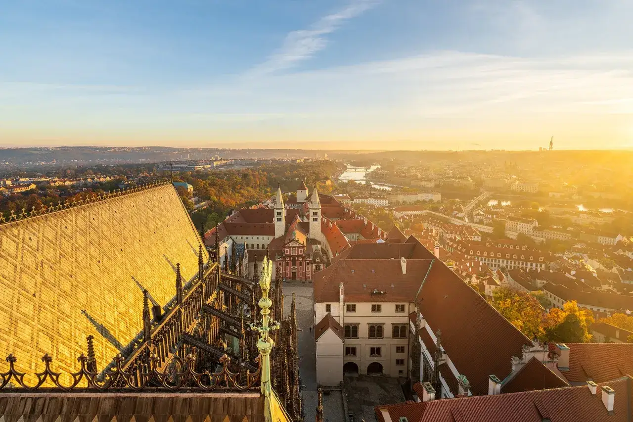 Vue panoramique du château de Prague baigné par le soleil levant, avec ses toits de tuiles rouges et la ville s'étendant à perte de vue.