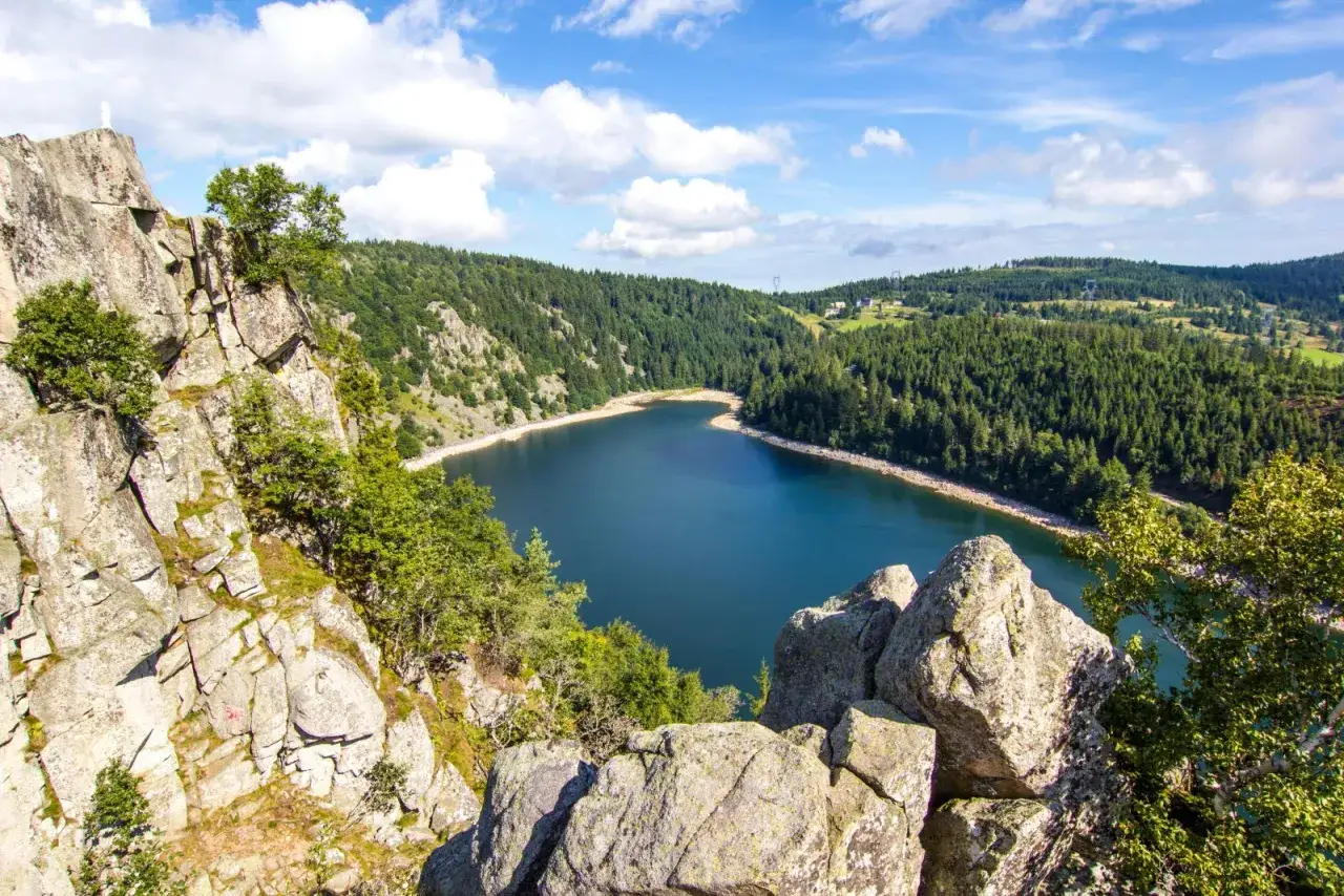 Vue panoramique d'un lac bleu entouré de forêts verdoyantes, avec des rochers imposants au premier plan. Parfait pour des vacances dans les Vosges.