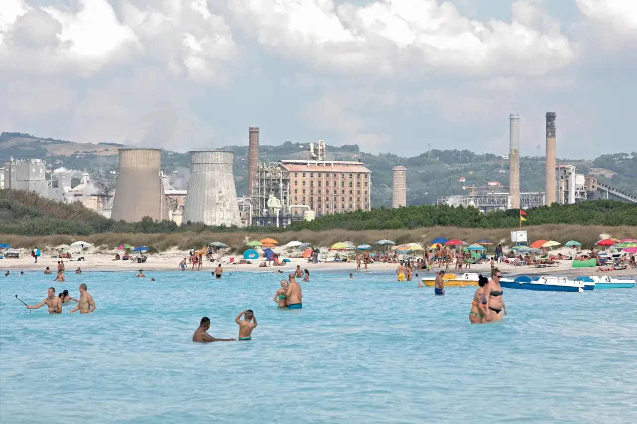 Plage animée à Rosignano Marittimo, avec des baigneurs dans l'eau turquoise et des installations industrielles en arrière-plan.