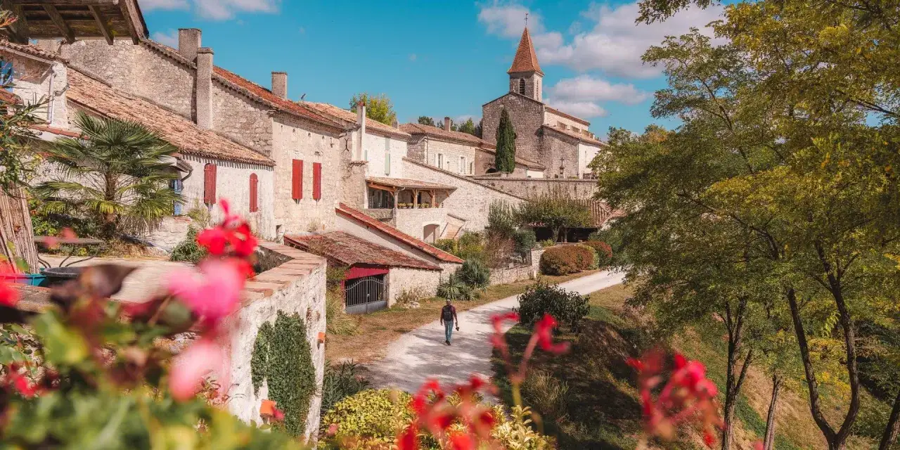 Un homme marche dans un village pittoresque, parmi les plus beaux villages du Tarn-et-Garonne. L'église et ses maisons en pierre se dressent sous un ciel bleu.