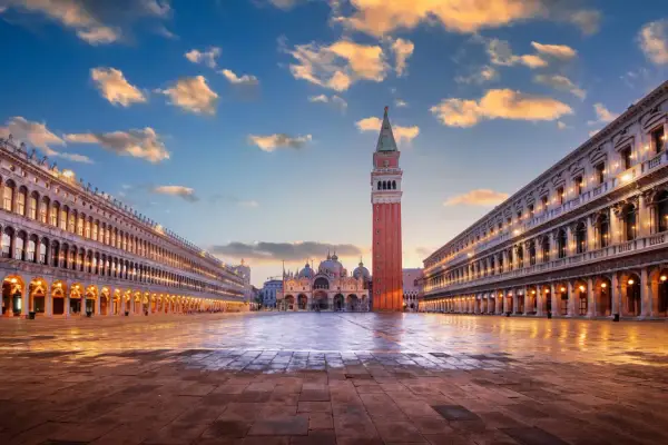 La piazza San Marco s'éveille sous un ciel aux teintes orangées. La basilique et le campanile se dressent majestueusement, reflétés sur le sol mouillé.