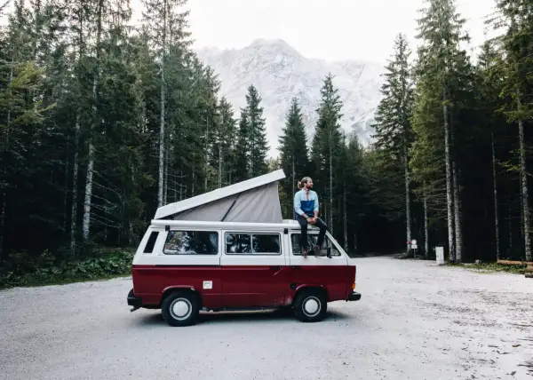 Un homme profite d'une pause lors de son tour d'Europe en van, assis sur le toit, entouré d'une forêt de pins et de montagnes majestueuses.