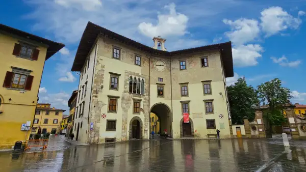 La Piazza dei Cavalieri à Pise, avec ses bâtiments historiques aux façades ocre et jaune, sous un ciel bleu et nuageux. Le sol mouillé reflète les édifices.