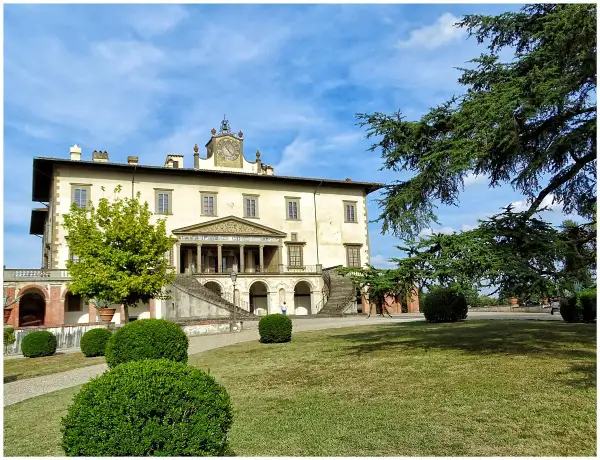 La villa Poggio a Caiano, un joyau architectural, se dresse sous un ciel bleu. Des arbres majestueux ombragent la pelouse verdoyante et les buissons taillés.