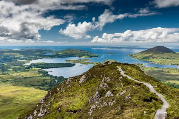 Vue panoramique du Connemara depuis Galway. Un sentier serpente sur une colline verdoyante, menant à des lacs bleus et des îles sous un ciel nuageux.