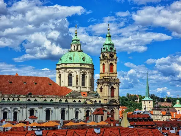 Vue imprenable sur l'église Saint-Nicolas de Prague, avec ses dômes verts et sa tour d'horloge, sous un ciel bleu et nuageux.