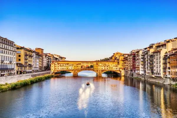 Le Ponte Vecchio, avec ses boutiques colorées, enjambe l'Arno sous un ciel bleu. Une petite barque glisse sur l'eau.