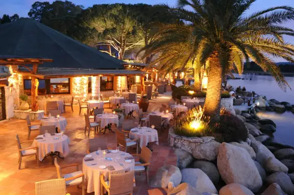 Terrasse illuminée d'un restaurant pieds dans l'eau, palmiers et tables dressées. L'endroit idéal pour un dîner au meilleur restaurant Porto Vecchio.