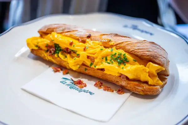 Sandwich aux œufs brouillés et bacon, parfait pour savoir où manger à Florence. Servi sur une assiette blanche avec une serviette "La Ménagère".
