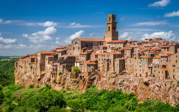 Vue panoramique de Magliano in Toscana, un village médiéval perché sur une falaise, avec ses maisons en pierre et son clocher imposant sous un ciel bleu.