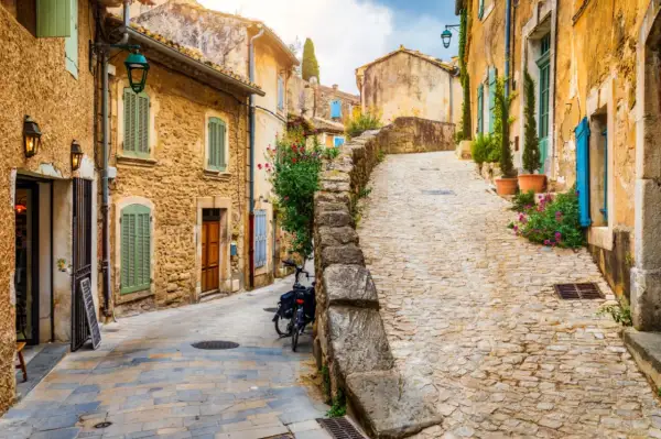 Ruelle pavée d'un village du Luberon, maisons en pierre ocre, volets verts et bleus, fleurs colorées. Parfait pour un week-end Luberon.