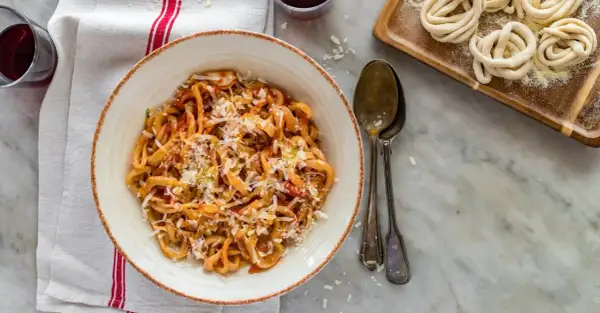 Pici all'aglione, pâtes toscanes savoureuses, servies dans un bol avec du fromage râpé. À côté, des pâtes crues enroulées.