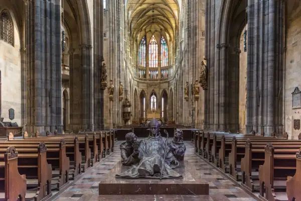 Intérieur de la cathédrale Saint-Guy au **château de Prague**. Une sculpture de bronze d'un évêque et deux moines trône au centre, face aux bancs de bois et aux vitraux colorés.